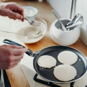 Close-up of pancakes cooking in a pan on a gas stove with utensils nearby.